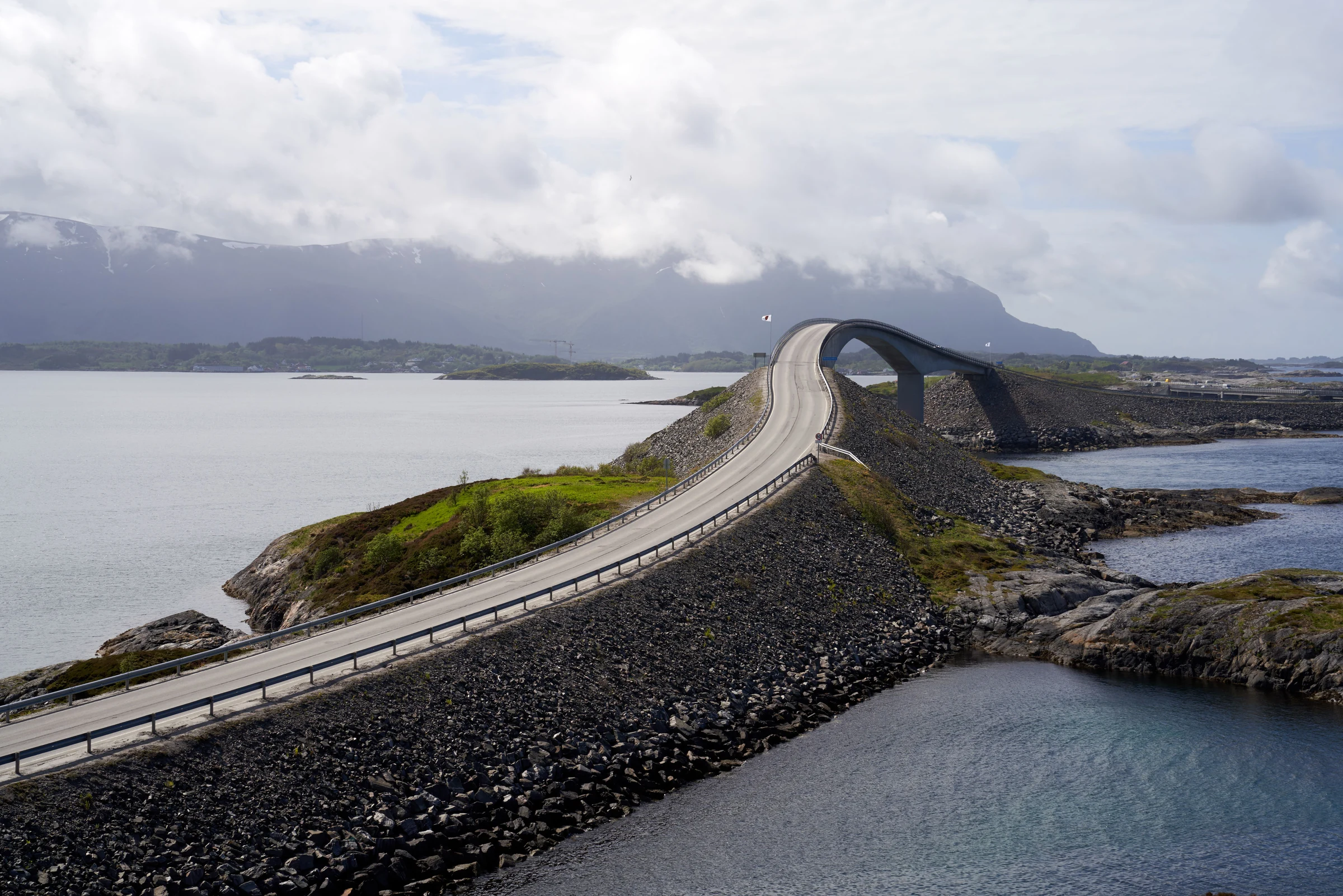A sweeping road bridge between sea and mountains beneath a clouded sky.