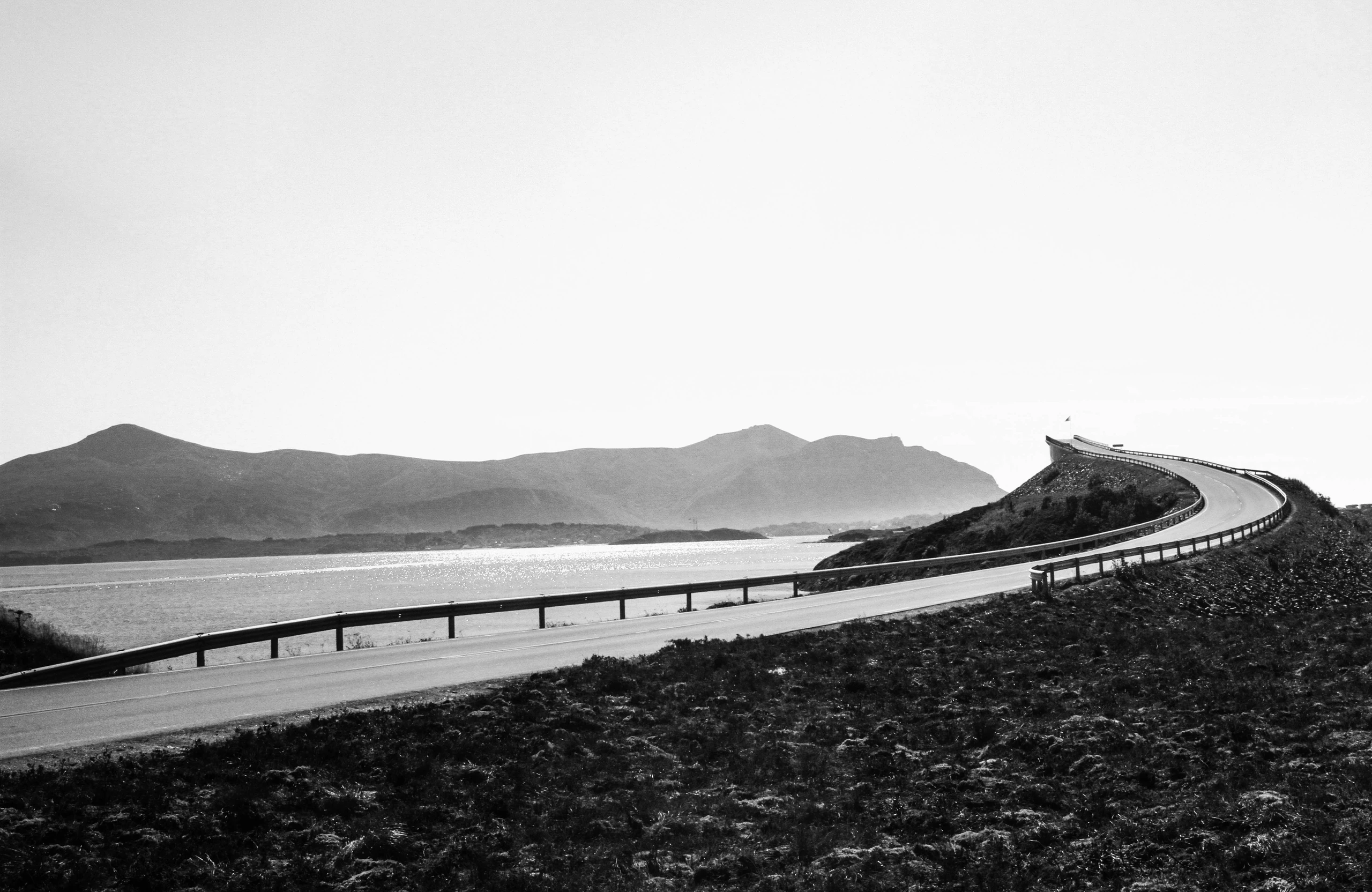 An empty coastal road curving towards the horizon, photographed in black and white.